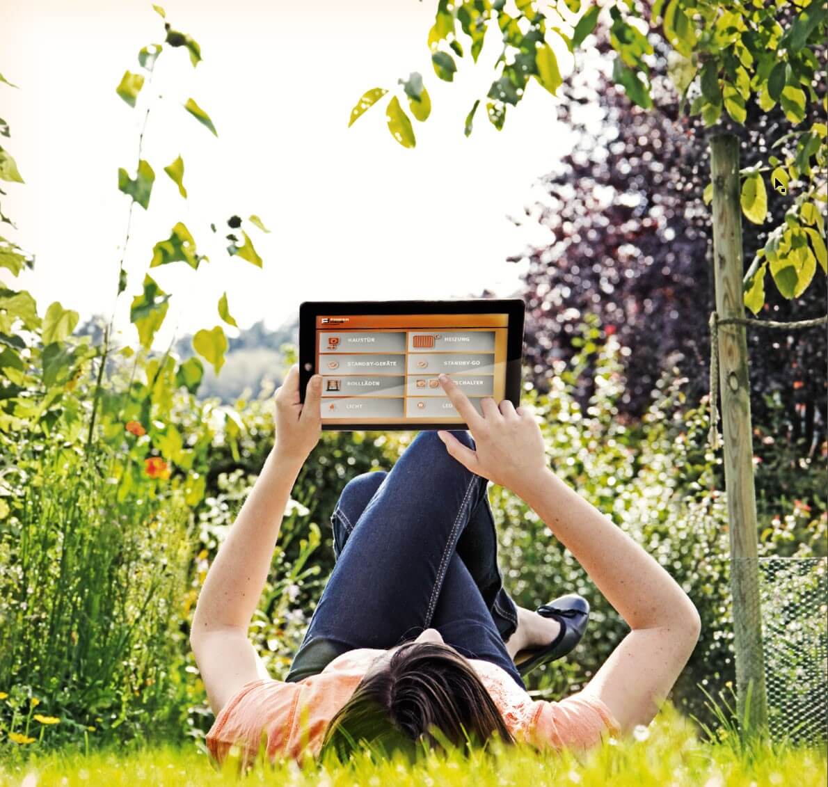 Frau liegt im Gras und hat ein Tablett in der Hand mit dem sie ihr Finger-Haus steuern kann.