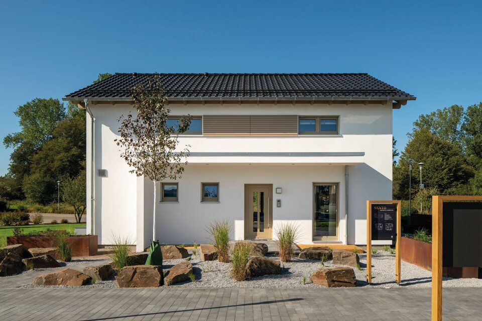 Frontansicht des Finger-Haus Musterhauses SENTO 500 in Frankenberg mit weißer Putzfassade, dunkel gedecktem Satteldach und horizontalem Fassadenelement; davor gepflasterter Weg, Kiesfläche mit Natursteinen, Ziergräsern, kleinem Baum und Informationsstele.