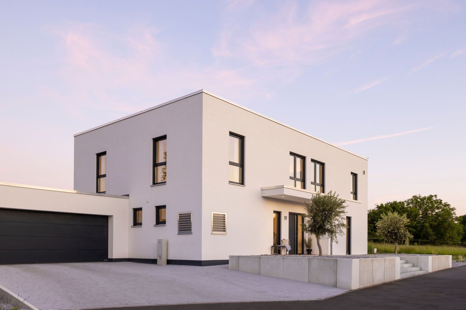 Modernes FingerHaus im kubischen Bauhausstil mit weißer Fassade, Flachdach, großer Garage und gepflasterter Einfahrt, aufgenommen bei Abendlicht.