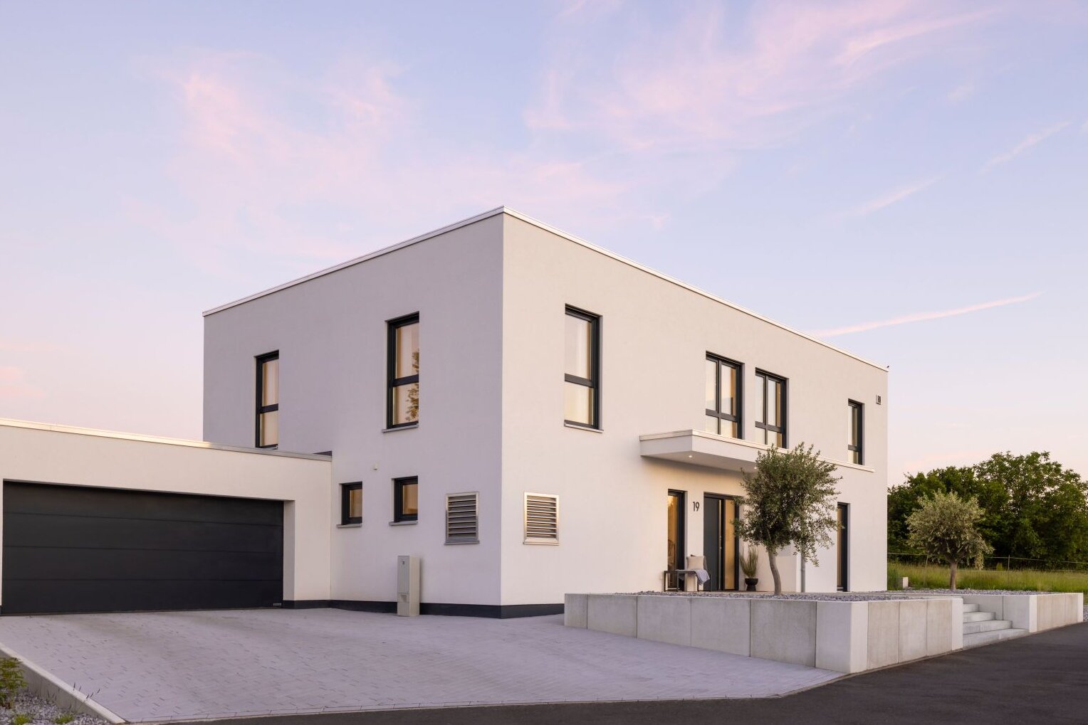 Modernes FingerHaus im kubischen Bauhausstil mit weißer Fassade, Flachdach, großer Garage und gepflasterter Einfahrt, aufgenommen bei Abendlicht.