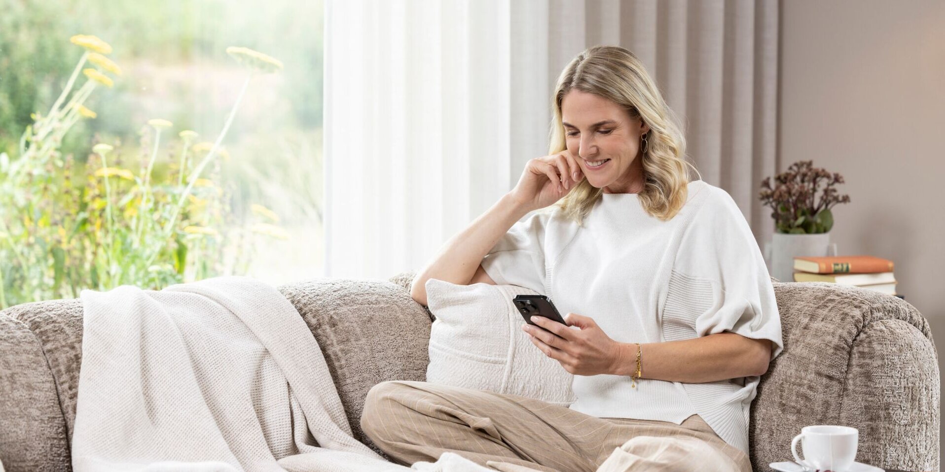 Finger-Haus-Besitzerin sitzt entspannt auf einem Sofa im Wohnraum und blickt lächelnd auf ein Smartphone; neben ihr liegen eine Decke und Kissen, daneben ein Beistelltisch mit Kaffeetasse und Buch, im Hintergrund bodentiefes Fenster mit Vorhang und Gartenblick.