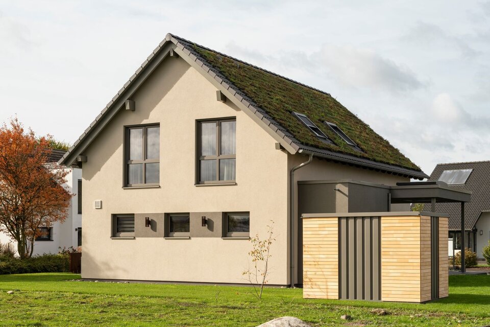 Seitenansicht des Musterhauses JUNO 300 in Mülheim-Kärlich mit begrüntem Dach, großen Fenstern und moderner Holzverkleidung.