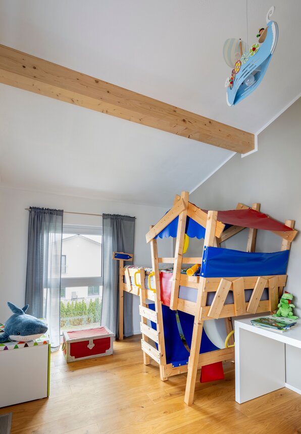 Kinderzimmer mit hölzernem Hochbett in Hausform, blauen und roten Stoffelementen, großem Fenster und Holzboden.
