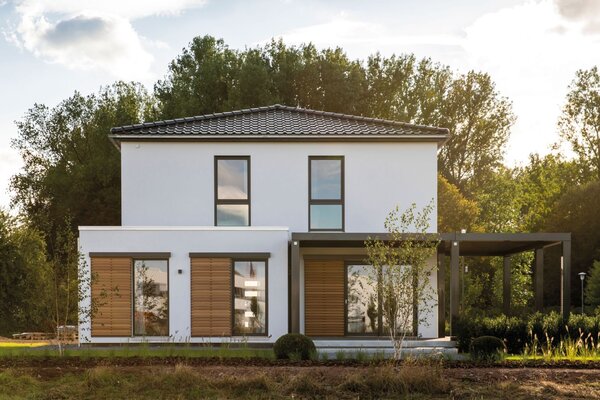 Frontansicht des Musterhauses MEDLEY 300 B in Frankenberg mit klarer Architektur, Holzlamellen und seitlicher Pergola bei Sonnenuntergang.