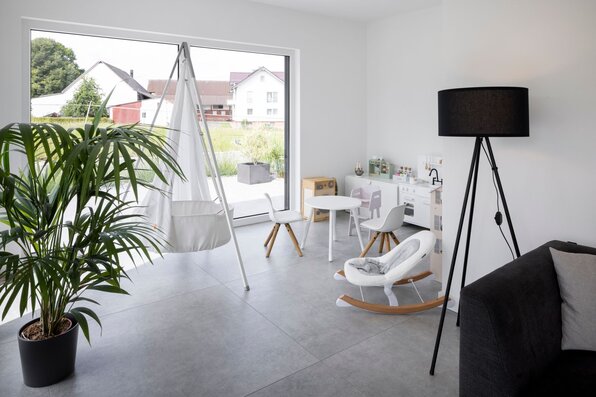 Modernes Spielzimmer mit großer Fensterfront, Hängesitz, Kindertisch und Spielküche in einem frei geplanten FingerHaus.