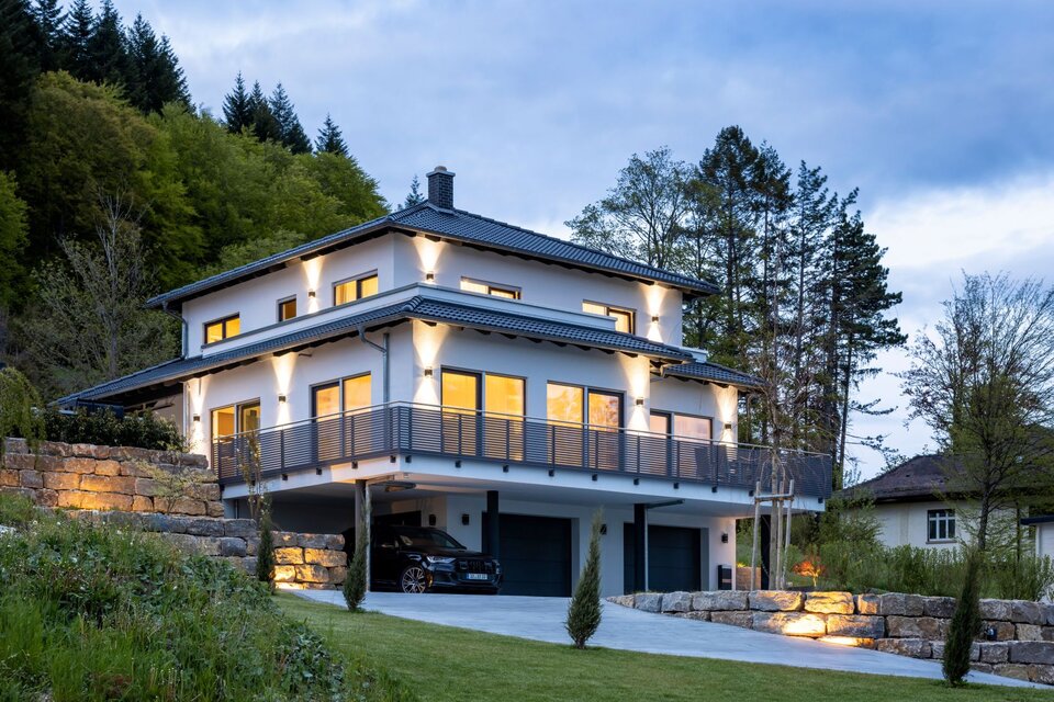 Modernes FingerHaus mit Walmdach und umlaufendem Balkon, stimmungsvoll beleuchtet in der Abenddämmerung.