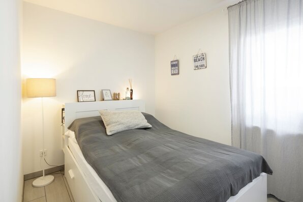 Schlafzimmer im SENTO 403 mit weißem Bett, grauer Tagesdecke, Stehleuchte und Fenster mit Vorhang