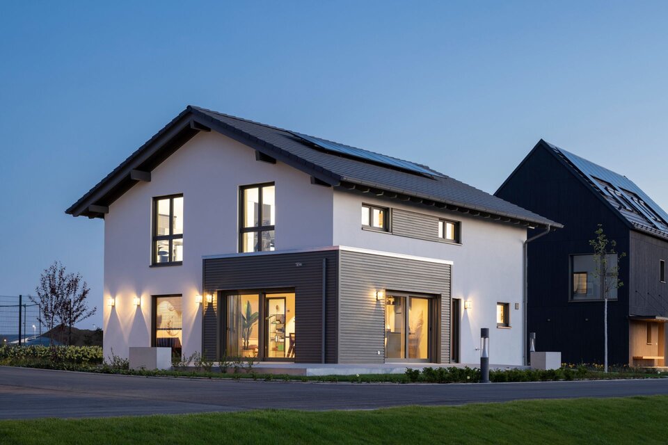 FingerHaus ARTIS 400 Musterhaus im Schwarzwald bei Abenddämmerung mit eingeschalteter Außenbeleuchtung, Satteldach, Photovoltaikanlage und Holz-Erker.