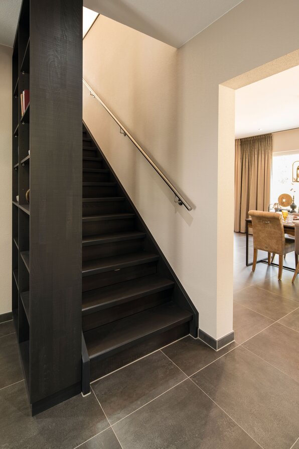 Schwarze Treppe mit Edelstahlhandlauf neben offener Durchgangstür zu einem Esszimmer mit Tisch und Stühlen