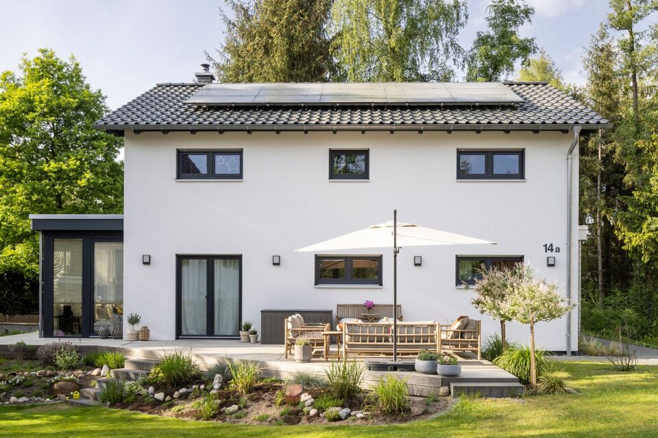 Modernes frei geplantes FingerHaus mit heller Putzfassade, großer Terrasse und gepflegtem Garten, umgeben von Bäumen und Natur.
