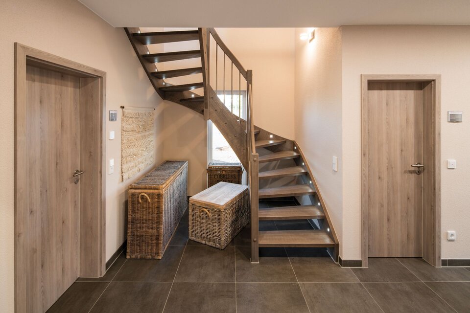Eingangsbereich mit dunklem Fliesenboden, offene Holztreppe mit Metallgeländer und zwei Rattantruhen unter der Treppe. Helle Wände, Holztüren und dezente Beleuchtung schaffen ein warmes Ambiente im Musterhaus UNO in Leipzig.