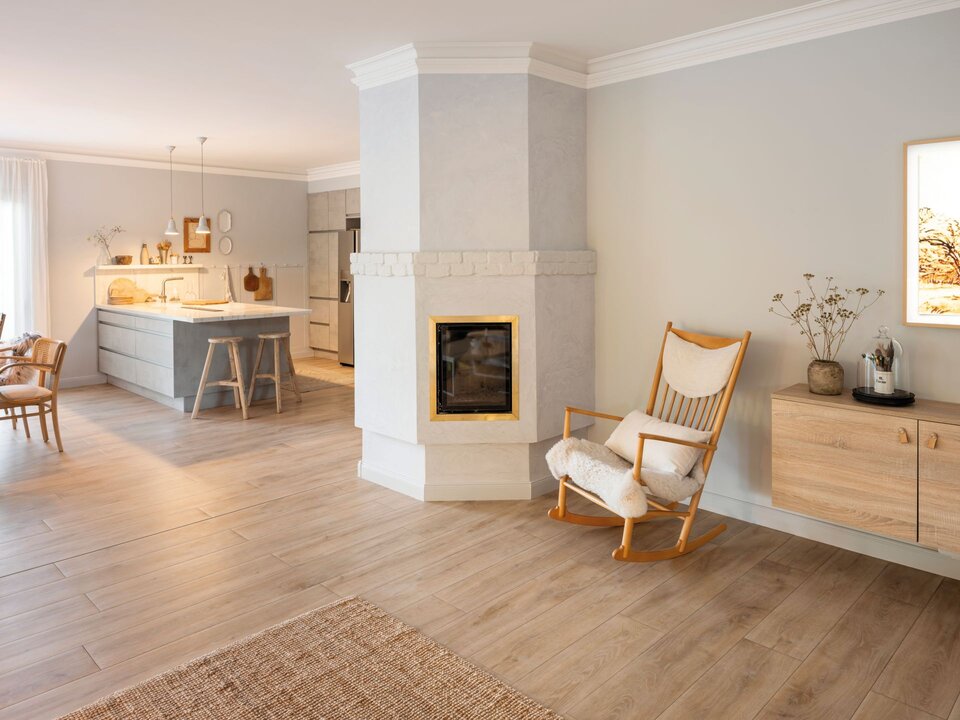 Wohnbereich mit Kamin, Holzfußboden und hellen Wänden. Ein gemütlicher Sessel, Sideboard und angrenzende offene Küche schaffen eine warme, freundliche Atmosphäre.