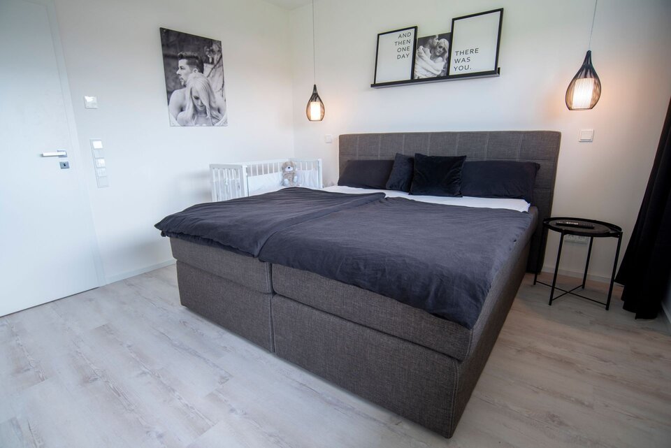 Schlafzimmer in einem FingerHaus NIVO 130 mit großem Boxspringbett in dunklen Farbtönen, Wandbildern, zwei modernen Hängelampen und einem Babybeistellbett.