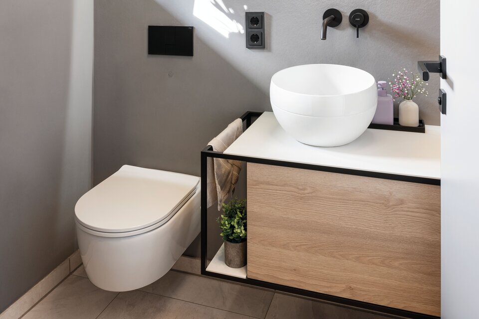 Modernes Bad mit Wand-WC, rundem Spiegel und weißem Aufsatzwaschbecken auf Holzunterschrank. Schwarze Armaturen und helle Bodenfliesen ergänzen das schlichte, elegante Design.