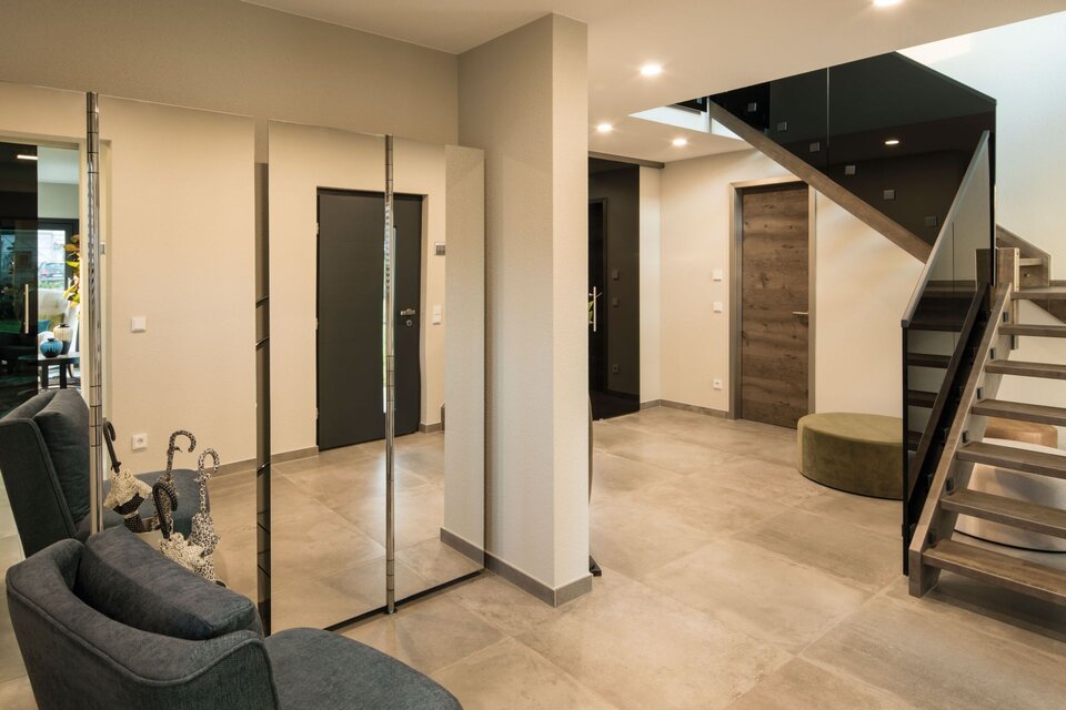 Moderner Eingangsbereich eines FingerHaus Fertighauses mit grauem Sofa, großem Spiegel, hellen Fliesenboden, Holztreppe und mehreren Türen