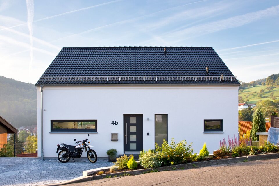 Modernes FingerHaus-Einfamilienhaus mit weißer Putzfassade, dunklem Satteldach, klarer Linienführung und Vorgarten, gelegen in Hanglage mit Motorrad in der Einfahrt.