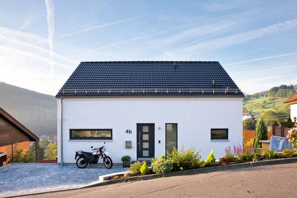 Modernes FingerHaus-Einfamilienhaus mit weißer Putzfassade, dunklem Satteldach, klarer Linienführung und Vorgarten, gelegen in Hanglage mit Motorrad in der Einfahrt.