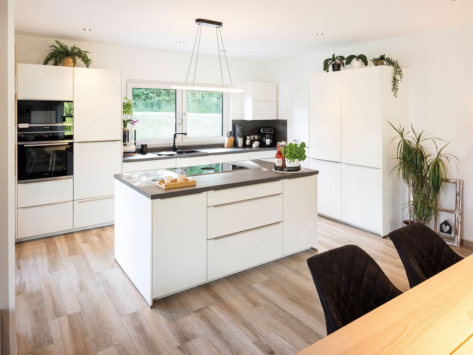 Moderne Küche in Weiß mit großer Kochinsel, dunkler Arbeitsplatte und Essbereich im Vordergrund. Pflanzen, Pendelleuchten und Holzfußboden betonen das helle Ambiente.