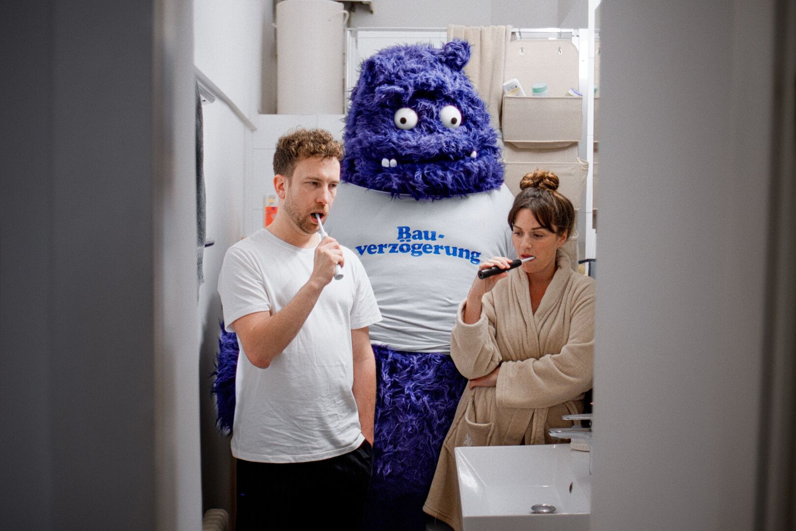 Ein Paar steht im Badezimmer und putzt sich die Zähne, während hinter ihnen ein großes blaues Monster mit der Aufschrift „Bauverzögerung“ auftaucht – Teil der FingerHaus-Kampagne „Das Unerwartete“.