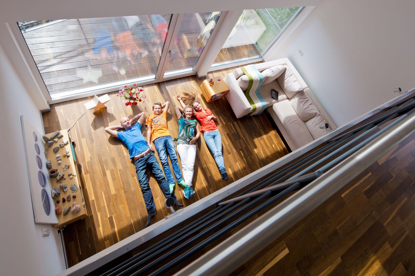 Familie liegt entspannt auf dem Holzboden des hellen Wohnzimmers mit Galerie und großen Panoramafenstern.
