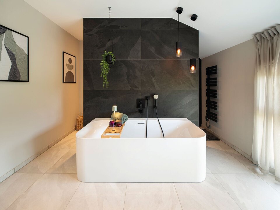 Modernes Badezimmer mit freistehender weißer Badewanne vor dunkler T-Wand, zwei schwarzen Pendelleuchten und hellen Bodenfliesen. An der Wand hängt ein schwarzer Handtuchheizkörper, daneben Fenster mit Vorhängen und Wandbilder.