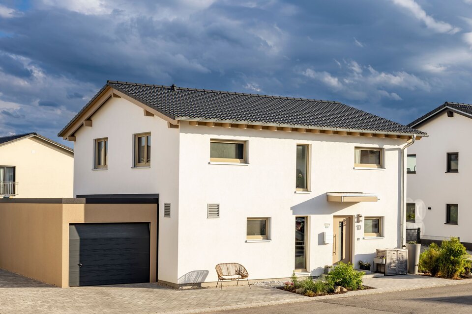 Zweigeschossiges weißes Einfamilienhaus mit dunklem Satteldach, angebauter Garage und gepflastertem Vorplatz.