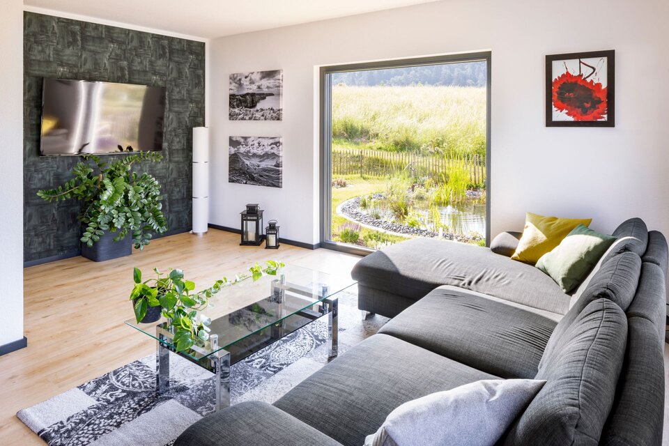 Wohnzimmer mit grauer Couch, Couchtisch und dunkler Akzentwand mit Fernseher. Große Fenster und Pflanzen ergänzen das moderne Ambiente.