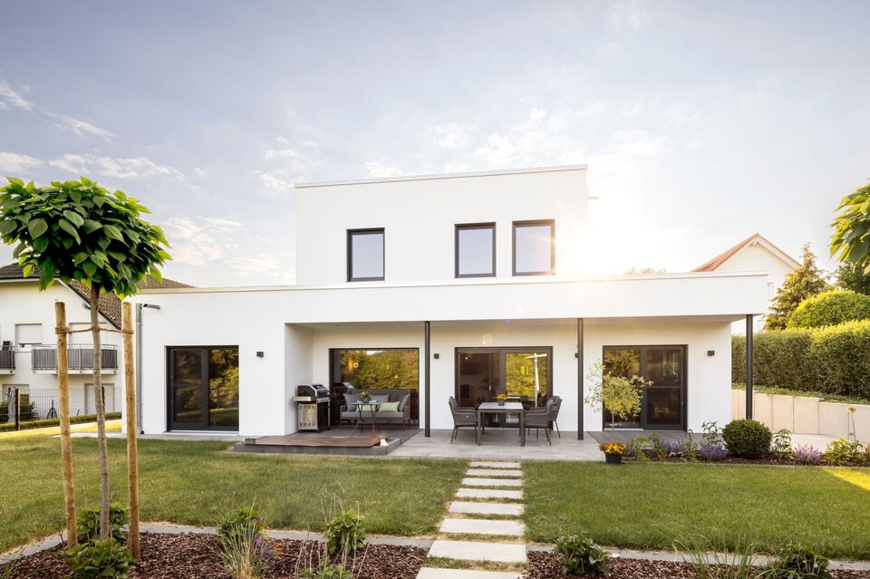 Moderne Außenansicht eines frei geplanten FingerHaus Einfamilienhauses mit weiß verputzter Fassade im kubischen Bauhaus-Stil. Mit überdachter Terrasse, großen Fenstern und gepflegtem Garten.