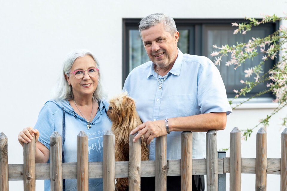 Paar mit kleinem Hund steht lächelnd hinter einem Holzzaun vor einem Haus