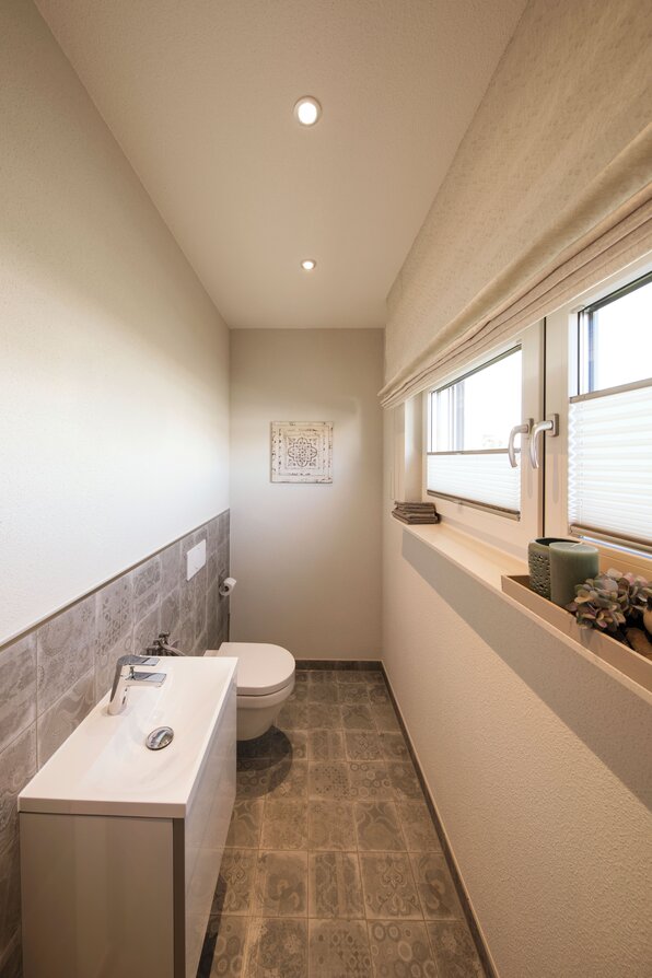 Modernes Gäste-WC im FingerHaus NEO Musterhaus Günzburg mit hellem Waschtisch, Fenster und stilvollen Fliesen.