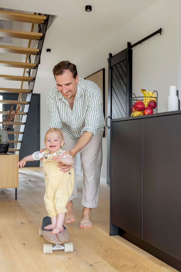 Ein Vater spielt mit seinem kleinen Kind im modernen Wohnbereich eines FingerHaus-Hauses. Das Kind steht lachend auf einem Skateboard, während der Vater es festhält.