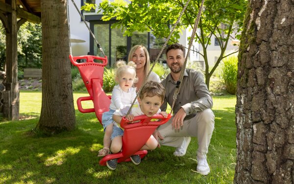 Familie genießt den sonnigen Garten eines frei geplanten FingerHaus – zwei Kinder schaukeln unter Bäumen, während die Eltern daneben sitzen und lächeln.