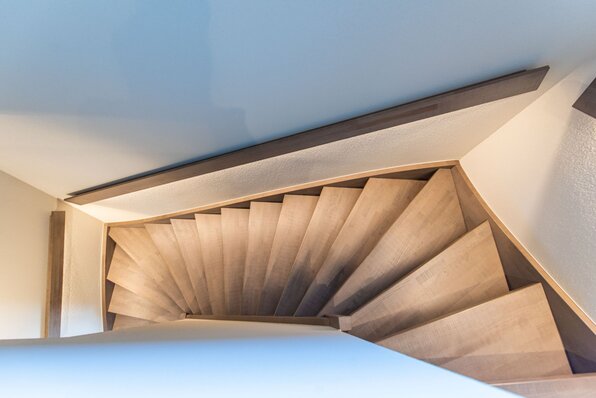 Holztreppe mit gleichmäßig verlaufenden Stufen in einem modernen Treppenhaus im Musterhaus TALO Babenhausen. Helle Wände und klare Linien betonen das warme Holz und die geometrische Form.