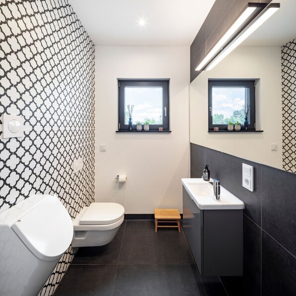 Modernes Badezimmer in einem FingerHaus-Haus mit stilvoller schwarz-weißer Fliesenwand, wandhängender Toilette, großem Spiegel und Fenster für natürliches Licht.