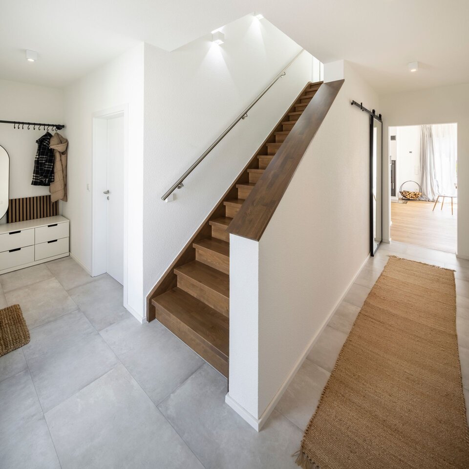 Heller Flur mit geschlossener gerader Holztreppe, weißem Geländer und grauem Boden. Eingangsbereich mit Garderobe, hellen Wänden und klarer Linienführung.