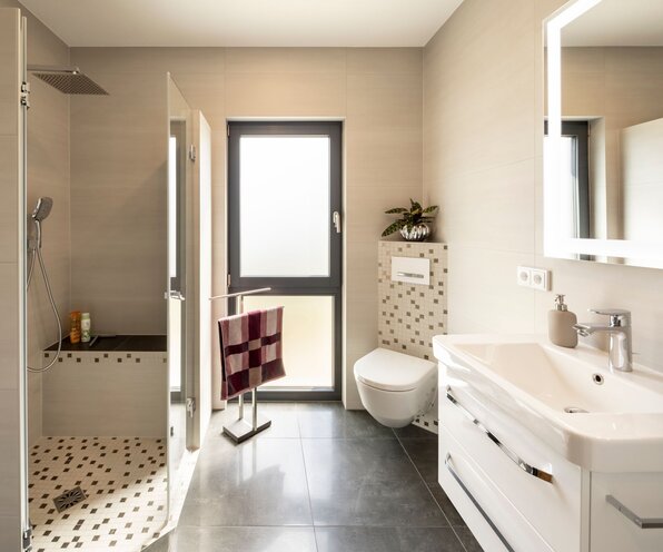 Modernes Badezimmer in einem frei geplanten FingerHaus mit bodentiefer Dusche, WC und großem Waschtisch mit Spiegel. Helle Fliesen kombiniert mit Mosaikakzenten und dunklem Boden schaffen ein elegantes Ambiente.