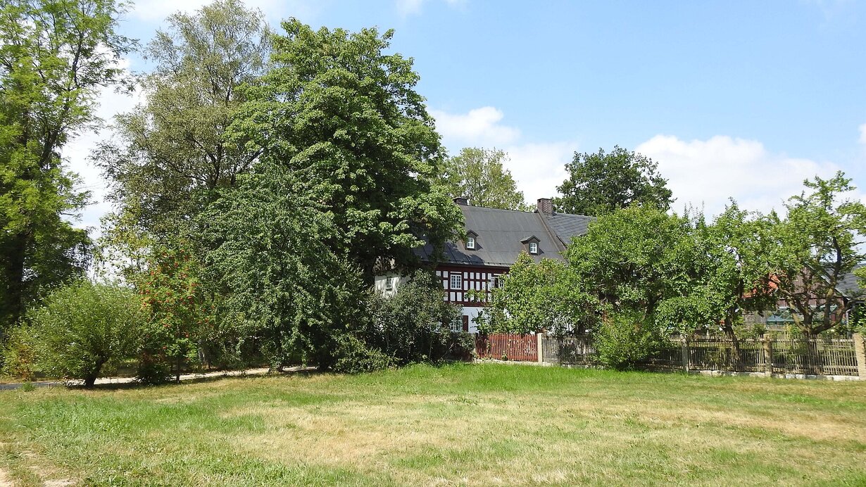 Traditionelles Wohnstallhaus mit Fachwerk in Tröstau, umgeben von hohen Bäumen und einer grünen Wiese unter blauem Himmel.