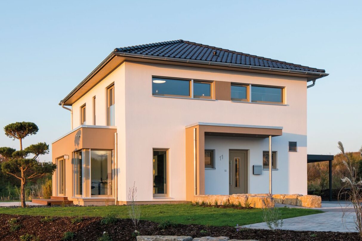 Außenansicht des Finger-Haus Musterhauses MEDLEY 300 B in Kassel: zweigeschossiges Haus mit weißer Fassade, dunkel gedecktem Walmdach und großem Eckfenster; davor Rasenfläche, Steinmauer aus Natursteinen, Mulchbeet und gepflasterte Fläche bei tief stehender Sonne.