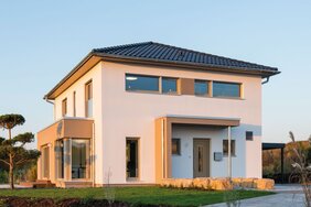 Außenansicht des Finger-Haus Musterhauses MEDLEY 300 B in Kassel: zweigeschossiges Haus mit weißer Fassade, dunkel gedecktem Walmdach und großem Eckfenster; davor Rasenfläche, Steinmauer aus Natursteinen, Mulchbeet und gepflasterte Fläche bei tief stehender Sonne.