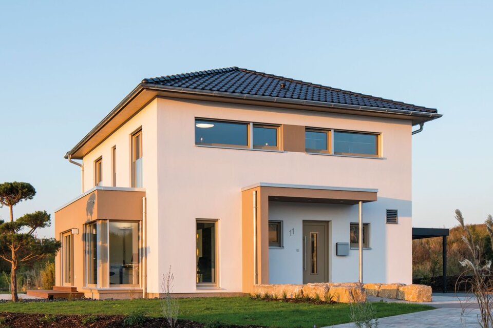 Außenansicht des Finger-Haus Musterhauses MEDLEY 300 B in Kassel: zweigeschossiges Haus mit weißer Fassade, dunkel gedecktem Walmdach und großem Eckfenster; davor Rasenfläche, Steinmauer aus Natursteinen, Mulchbeet und gepflasterte Fläche bei tief stehender Sonne.