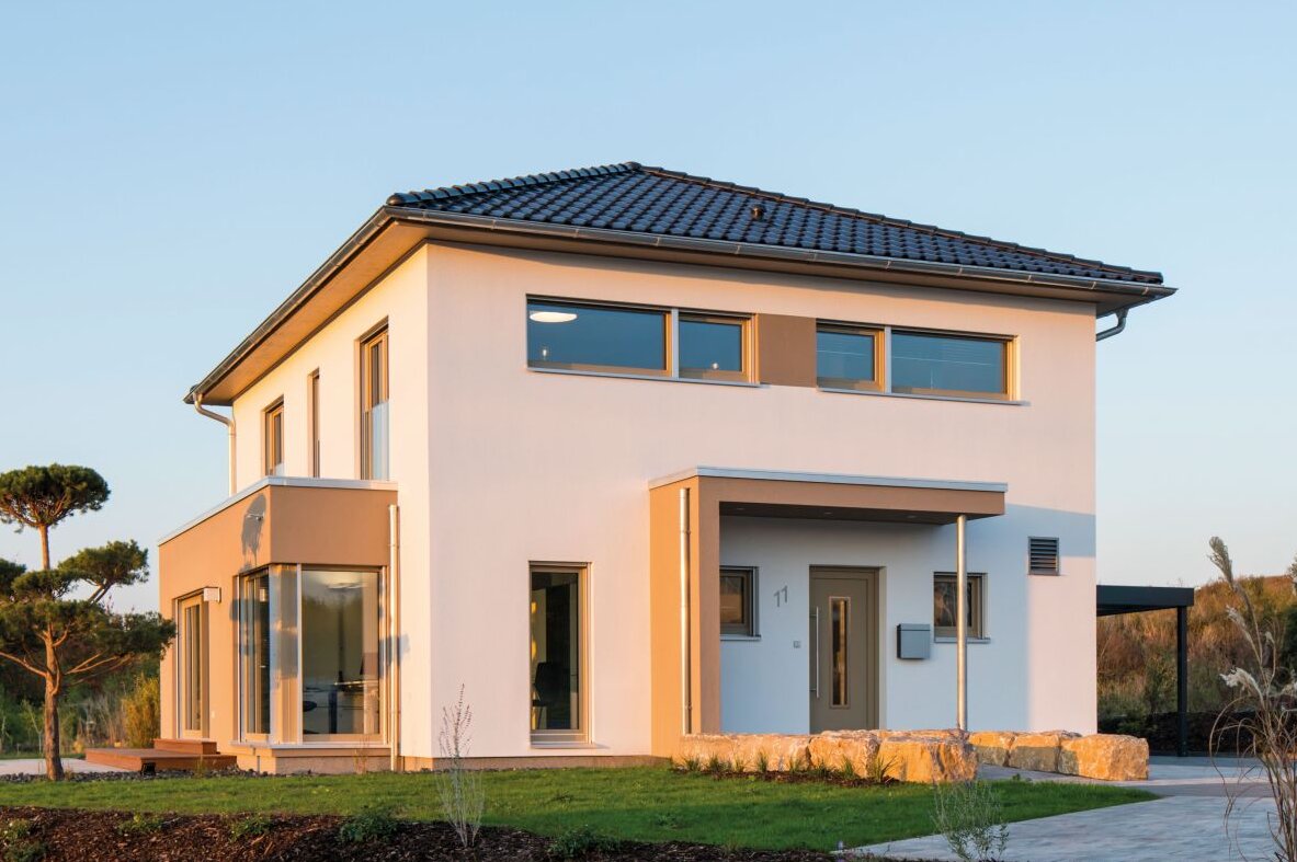 Außenansicht des Finger-Haus Musterhauses MEDLEY 300 B in Kassel: zweigeschossiges Haus mit weißer Fassade, dunkel gedecktem Walmdach und großem Eckfenster; davor Rasenfläche, Steinmauer aus Natursteinen, Mulchbeet und gepflasterte Fläche bei tief stehender Sonne.
