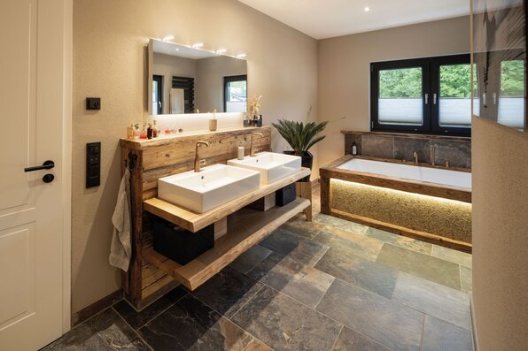 Modernes Badezimmer in einem FingerHaus-Haus mit Doppelwaschtisch aus rustikalem Holz, eingelassener Badewanne und stilvoller Beleuchtung.