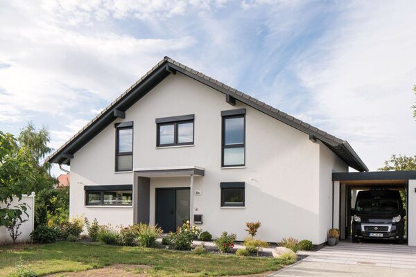 Frei geplantes Einfamilienhaus mit heller Fassade, Satteldach, Carport und gepflegtem Vorgarten, aufgenommen aus der Straßenperspektive.