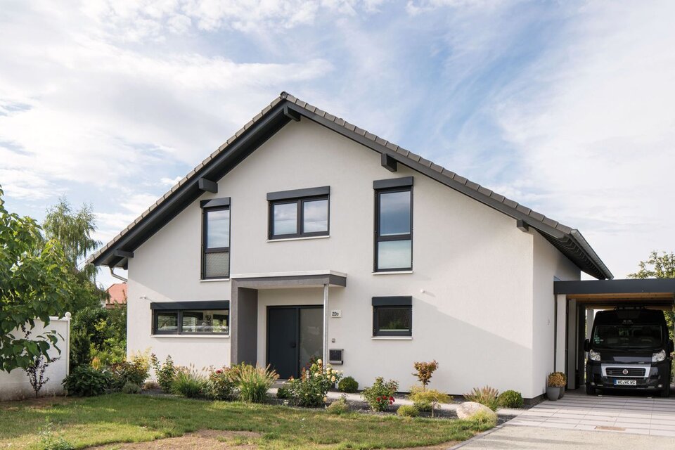 Frei geplantes Einfamilienhaus mit heller Fassade, Satteldach, Carport und gepflegtem Vorgarten, aufgenommen aus der Straßenperspektive.