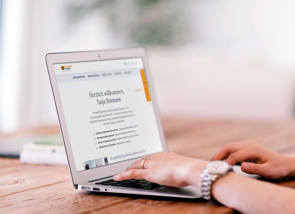 Person tippt auf einem Laptop am Holztisch, auf dem Bildschirm ist die FingerHaus Website mit persönlicher Begrüßung und Navigationsmenü zu sehen.
