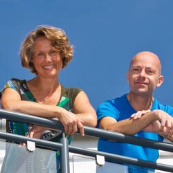 Vierköpfige Familie lehnt am Geländer ihres modernen Balkons und blickt fröhlich in die Kamera vor blauem Himmel.