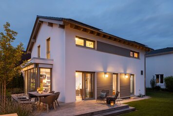 Stimmungsvolle Abendansicht des Musterhauses MEDLEY 300 B in Poing mit beleuchteter Terrasse und großen Fensterfronten.