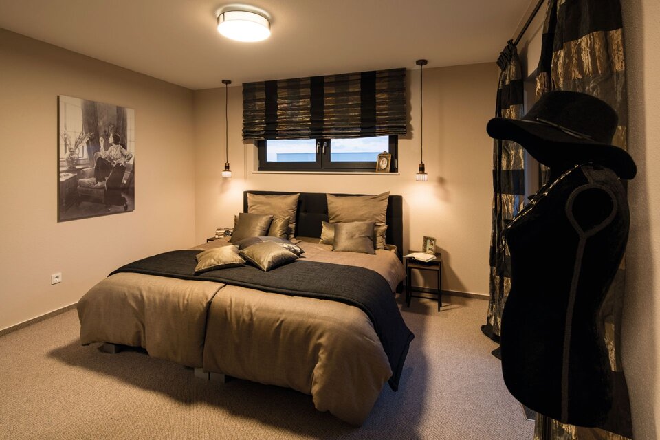 Schlafzimmer mit großem Bett, mehreren Kissen, einem Fenster mit Jalousien, einem stehenden Kleiderbügel mit Hut und einem schwarz-weißen Bild an der Wand