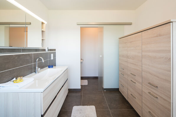 Modernes Badezimmer eines FingerHaus mit hellem Waschtisch, großem Spiegel und eleganten Holzfronten vor dunklen Fliesen.