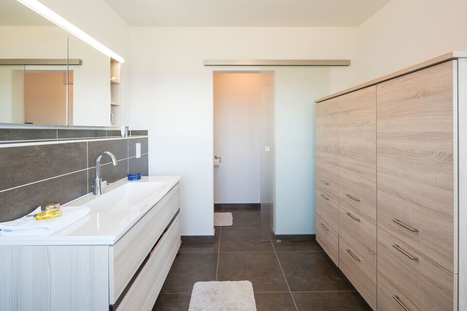 Modernes Badezimmer eines Finger-Hauses mit hellem Waschtisch, großem Spiegel und eleganten Holzfronten vor dunklen Fliesen.